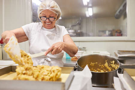Elderly female baker fills dough into mold while baking apple pie in bakery shopの写真素材