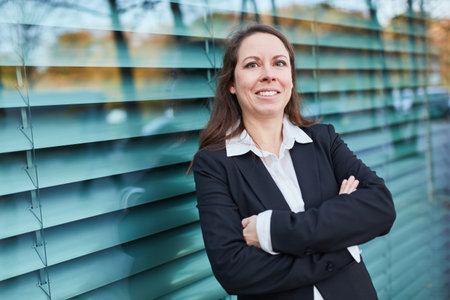 Smiling corporate businesswoman leaning outside the office on glass wall in cityの写真素材