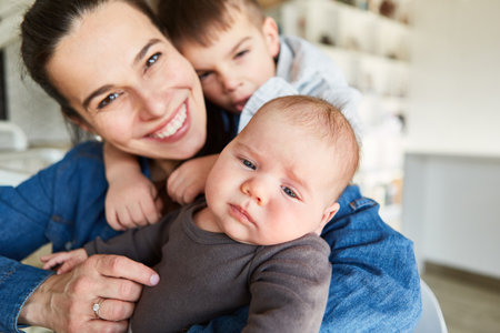 Happy smiling mother with baby and big brother cuddling together at homeの写真素材