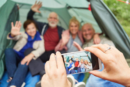Woman photographing senior couple through smart phone sitting in camping tent at forestの写真素材