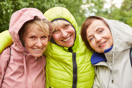 Portrait of smiling elderly female friends in jackets exploring forest during rainy seasonの写真素材