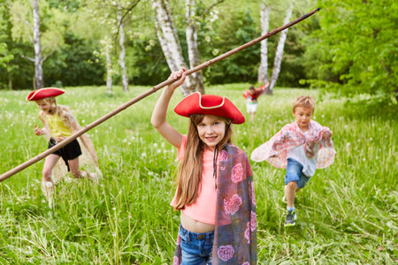 Portrait of smiling girl with tricorn hat and stick standing in grass by friends at parkの写真素材