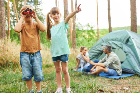 Boy looking through binoculars standing by sister pointing while camping in forest during vacationの写真素材