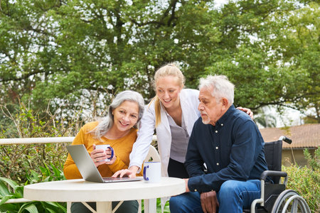 Smiling female caregiver discussing with elderly couple over laptop while having coffee in garden of nursing homeの写真素材