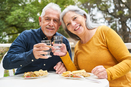 Portrait of happy senior man and woman toasting glass of water while having meal at table in gardenの写真素材