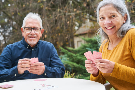 Portrait of senior man and woman playing cards while sitting at table in garden of nursing homeの写真素材