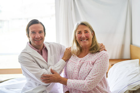 Portrait of smiling caring male doctor embracing elderly woman sitting on bed at rehabilitation...