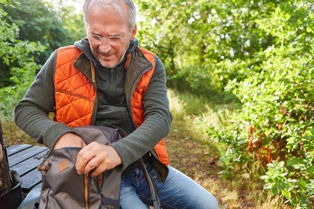 Senior man searching in backpack while sitting in forest during summer vacation