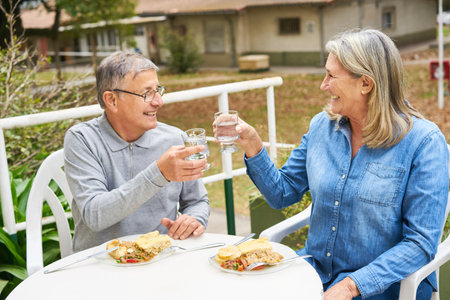 Cheerful senior man and woman toasting glass of water while having meal in garden of nursing home