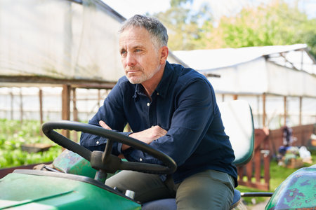 Thoughtful mature male farmer sitting with arms crossed on tractor by greenhouseの写真素材