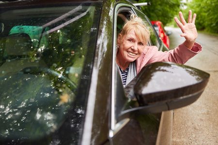 Portrait of cheerful senior man waving hand through windows while driving car during road tripの写真素材