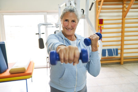 Portrait of smiling elderly woman exercising with dumbbells in rehabilitation center