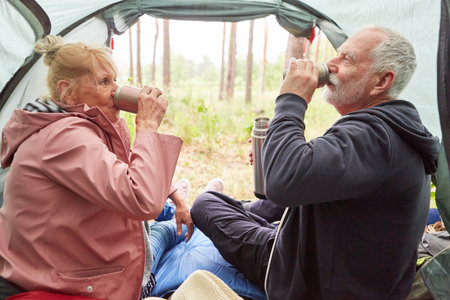 Elderly couple drinking tea together while relaxing in tent during camping at forestの写真素材