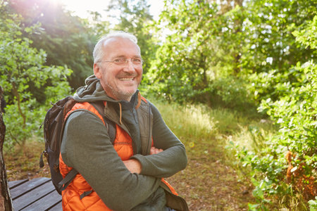 Portrait of happy senior man sitting with arms crossed in forest during summer vacationの写真素材