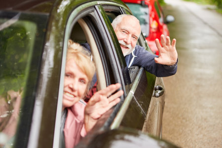 Portrait of happy elderly couple waving hands through windows while traveling in car during vacationの写真素材