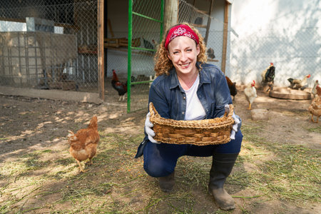 Portrait of smiling female farmer holding wicker basket crouching by hen at poultry farmの写真素材