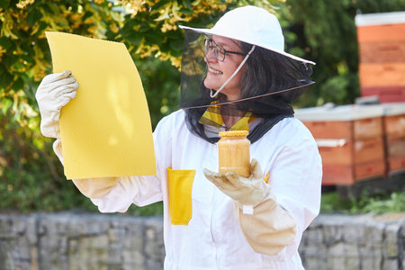 Female beekeeper offering honey jar and beeswax sheets in front of beehives during summerの写真素材