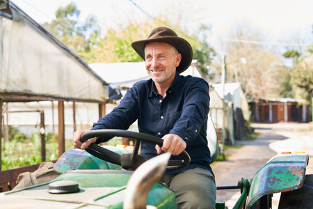 Happy mature male farmer in hat driving tractor by greenhouseの写真素材