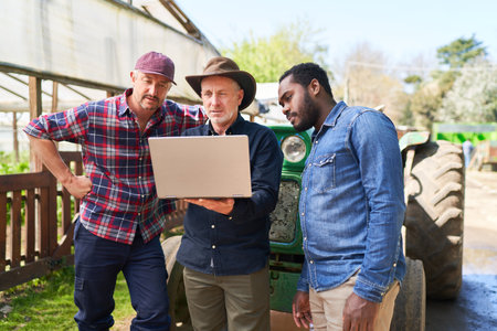 Mature male farmer discussing with colleagues over laptop in front of tractor at farmの写真素材