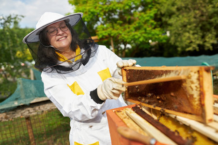 Happy senior female apiarist using brush on honeycomb frame at apiary garden