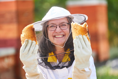 Portrait of happy female apiarist holding honeycomb wearing protective suit at apiary gardenの写真素材