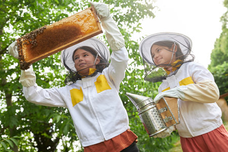 Girls with smoker examining honeycomb frame while wearing protective suit at apiary garden