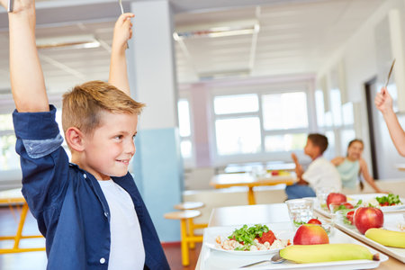 Smiling boy with arms raised sitting at table during lunch time in school cafeteriaの写真素材