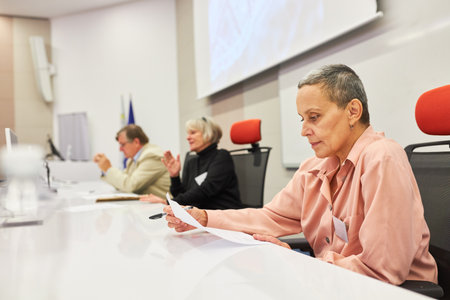 Mature female speaker reading document while sitting at panel in convention center