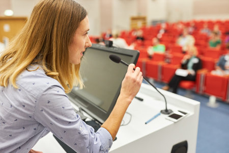 Female speaker on panel at business conference talking with audience through microphone