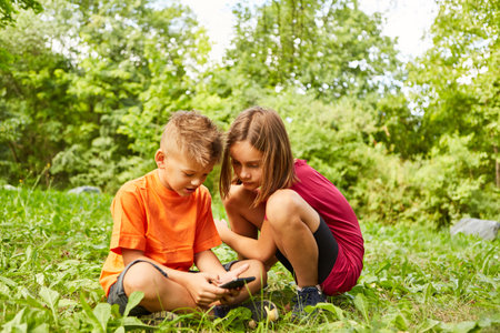 Girl crouching next to boy using smart phone while sitting on grass at parkの写真素材