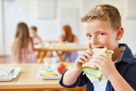 Portrait of hungry schoolboy eating fresh sandwich during lunch time at cafeteriaの写真素材