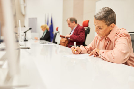 Mature female speaker writing in document while sitting at panel in convention centerの写真素材