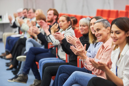 Smiling male and female professionals applauding while sitting in audience at business conference in...