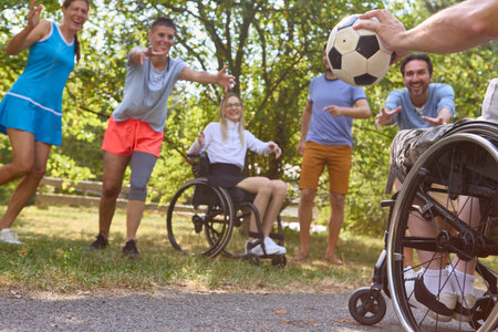 Group of friends, including a person in a wheelchair and others standing, enjoying a game of soccer in a sunny park, exuding joy and teamwork.の写真素材