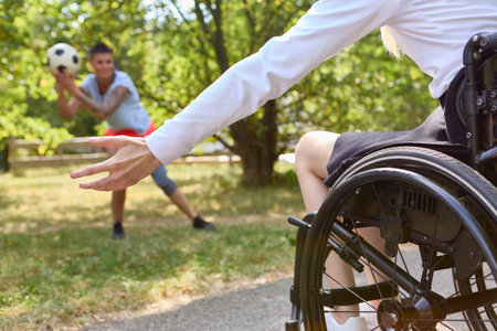 An active scene of a person in a wheelchair reaching out to play football with a friend in a lush, sunny park setting, showing inclusion and mobility.の写真素材