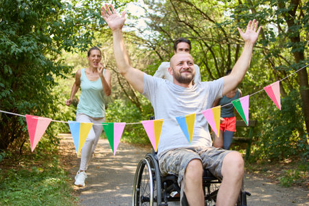 A joyful man who uses a wheelchair celebrates as he crosses a finish line, with supportive friends cheering in a park setting.の写真素材