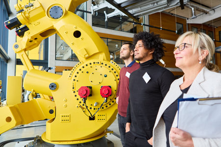 A diverse group of professionals observing a large yellow industrial robot arm in a modern facility,...