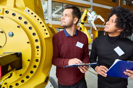 Two engineers reviewing and programming multi-purpose robotic arms in a modern industrial facility,...