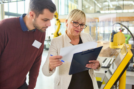 Two engineers reviewing documents and discussing robotics programming in an industrial setting,...