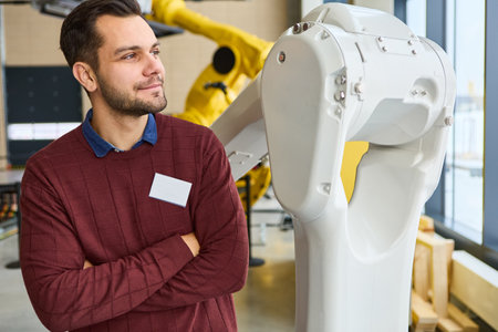 Engineer with folded arms standing next to a robotic arm in a modern industrial robotics lab,...