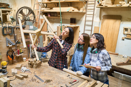 Three women engaged in a teamwork project at a lumberyard, showcasing skills and collaboration in a woodworking environment. The scene highlights mentorship and craftsmanship amidst stacks of wood and tools.の写真素材