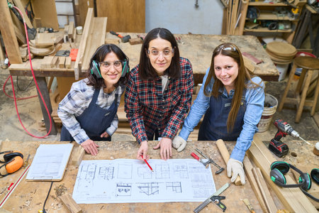 Three women collaborate on a woodworking project, examining blueprints in a lumberyard workshop. The scene highlights teamwork, skill development, and the importance of collaboration in craftsmanship.の写真素材