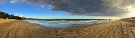 Wide beach panorama showcasing contrasting weather conditions with bright sun and dark clouds.の写真素材