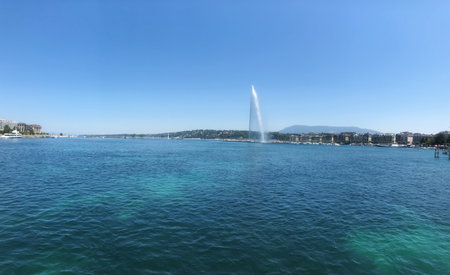 Wide-angle scene of a picturesque lake featuring a tall fountain, scenic surroundings, and clear skies, showcasing tranquility and natural beauty.の写真素材
