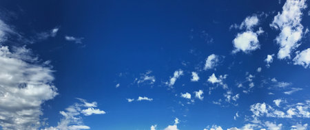 A wide panoramic view of a vibrant blue sky with scattered white fluffy clouds. The serene landscape...