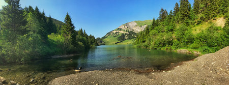 A panoramic view of a tranquil mountain lake surrounded by green forests and rocky peaks under a...