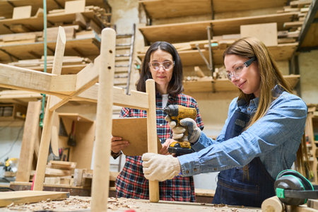 Two women in a lumberyard engage in woodworking as part of training and apprenticeship. They focus on teamwork and skill-building with a hands-on approach, highlighting professional and educational growth.の写真素材