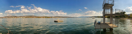A wide panoramic view of a peaceful lakeside, featuring a diving platform, clear calm waters, and a serene landscape ideal for tranquility and natural beauty appreciation.の写真素材