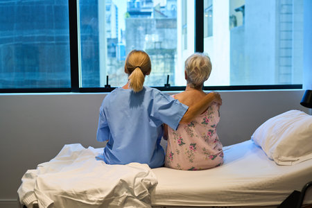 A nurse sits comforting an elderly woman in a hospital room during the daytime. The photo conveys care, support, and professional medical assistance in a serene healthcare environment.の写真素材