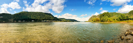 A wide panoramic view of a tranquil river surrounded by verdant hills and villages, capturing the essence of natural beauty and peacefulness with a backdrop of fluffy clouds and vibrant greenery.の写真素材